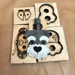 Peut inclure: Un porte-clés en cuir gris et blanc représentant une tête de Schnauzer, posé sur plusieurs planches de découpe en bois avec des lames en métal. Les planches en bois portent des inscriptions incluant 5-3, 5-2 et 5.