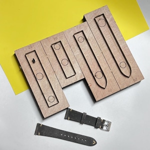May include: A set of brown wooden cutting dies for watch straps, featuring various rectangular and tapered shapes with circular cutouts and the number 20 engraved on the surface. Below the dies are two dark gray leather watch strap pieces.