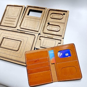 May include: A handmade, open, brown leather wallet with multiple card slots and a clear ID window. A blue credit card is visible in the ID window. Several wooden wallet pattern templates are in the background.