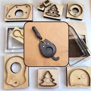 May include: A dark brown leather cat-shaped keyring with a circular cutout, a gold-tone metal swivel clasp, and a leather strap. The keyring is centred against a background of various wooden cutting dies used for leather crafting projects.