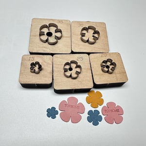 Könnte beinhalten: Eine Sammlung von lederverarbeitenden Stanzformen in Blumenform in verschiedenen Größen, beschriftet mit 15, 20, 25, 30 und 35. Die Stanzformen sind aus Metall und in Holzblöcken eingesetzt. Mehrere ausgeschnittene Lederblumen in Rosa, Blau und Gelb sind ebenfalls vorhanden.