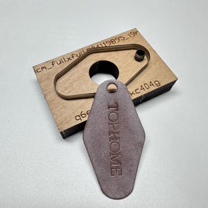 Puede incluir: Una etiqueta de llave de cuero marrón con el texto "TOP HOME" está al lado de un troquel de madera. El troquel tiene la forma de una etiqueta de llave con un agujero y un recorte circular. El troquel tiene el texto "kc4049" y otro texto.