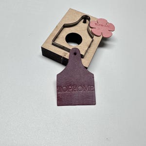 Könnte beinhalten: Ein mehrschichtiges Holzstanzteil mit Hausform, ein weinrotes Lederetikett und eine rosa Blume, alle mit dem Text "TOPHOME". Das Etikett befindet sich im Vordergrund, dahinter das Stanzteil und die Blume. Die Artikel liegen auf einer weißen Oberfläche.