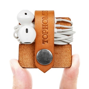 May include: A brown leather cord organiser with the word "TOPHOME" embossed on the front. The organiser has a metal button closure and is holding a pair of white earbuds and a tangle of white cords.