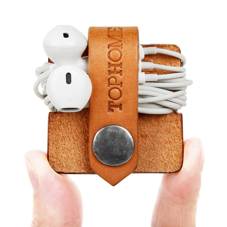 May include: A brown leather cord organiser with the word "TOPHOME" embossed on the front. The organiser has a metal button closure and is holding a pair of white earbuds and a tangle of white cords.