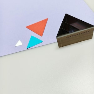 May include: A wooden triangle-shaped stamp with the word "STRAIGHT" printed on the side. There are four colorful paper triangles in red, blue, white, and orange. The objects are on a light purple surface.