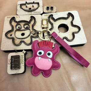 Peut inclure: Un porte-clés en forme de monstre magenta avec un ventre rose, des dents blanches et des cheveux rouges, attaché à un fermoir en métal doré et une dragonne assortie. Plusieurs moules de découpe en bois et métal avec des étiquettes BBD02 5/1.
