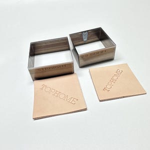May include: Two square metal leathercraft tools with wooden-look sides, labeled "STRAIGHT" and "23.80MM". Two square pieces of tan leather are stamped with the word "TOPHOME". The tools and leather are on a white surface.