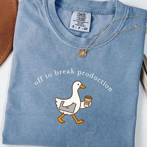 Puede incluir: Una camiseta de color azul vaquero con un gráfico de un ganso blanco que lleva un portátil y una taza de café. El texto sobre el ganso dice "off to break production" en letras blancas. La camiseta tiene cuello redondo y una etiqueta Comfort Colors.