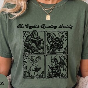 May include: A moss green t-shirt featuring the black text The Cryptid Reading Society above four square illustrations of a Bigfoot, a moth-like creature, a jackalope, and a dragon, each depicted reading a book within a leafy frame.