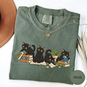 Puede incluir: Una camiseta de cuello redondo color verde musgo con un gráfico de cuatro gatos negros interactuando con pilas de libros y una pequeña planta en maceta. La prenda tiene una etiqueta Comfort Colors en el cuello y un collar de cadena dorada.