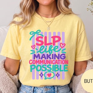 May include: A pale yellow short-sleeve t-shirt featuring a graphic print with vertical purple stripes and the text SLP life MAKING COMMUNICATION POSSIBLE in pink and teal, accented with hearts, a bow, and star shapes on the front center.