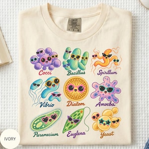 May include: An ivory t-shirt featuring a colorful graphic print of nine different microorganisms, each wearing sunglasses. The text labels include Cocci, Bacillus, Spirillum, Vibrio, Diatom, Amoeba, Paramecium, Euglena, and Yeast in various colors.