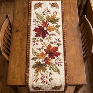 May include: A rectangular quilted table runner featuring a cream-colored background with a central print of autumn leaves, ferns, acorns, and berries. The runner is bordered by a brown and tan plaid fabric and includes cornucopia motifs.