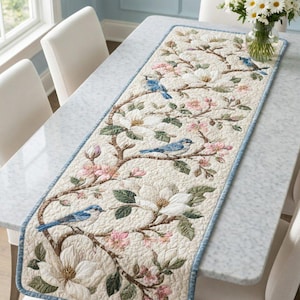 May include: A rectangular quilted table runner featuring a cream-colored background with a printed pattern of blue birds perched on brown branches among white magnolia flowers, pink blossoms, and green leaves, finished with a light blue border trim.