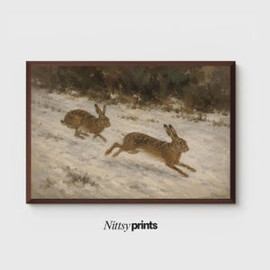 Puede incluir: Una impresión artística rectangular enmarcada que muestra una pintura de dos liebres marrones corriendo por un paisaje nevado con árboles oscuros al fondo. El texto Nittsy prints es visible en letras negras debajo de la obra enmarcada.