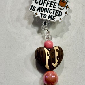 May include: A badge reel featuring a white acrylic topper with the black text MAYBE ICED COFFEE IS ADDICTED TO ME and an iced coffee graphic. Below are beads including a large brown heart, pink spheres, and a bead with a coffee cup pattern.