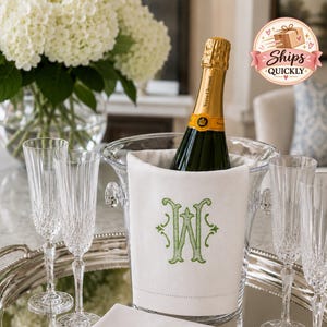 May include: A clear glass ice bucket containing a champagne bottle and a white linen wrap with a green embroidered letter W. The set includes four tall, fluted crystal glasses and a folded white linen napkin on a silver oval tray.