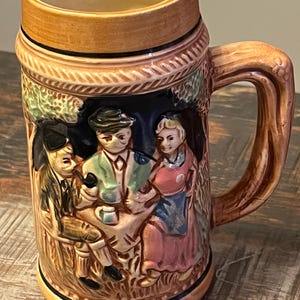 May include: A ceramic beer stein featuring a relief design of three figures sitting at a table. The mug has a tan rim and base, a brown handle, and a dark background. The figures are painted in muted tones of green, pink, and brown, with a rope-like texture around the top and bottom edges.