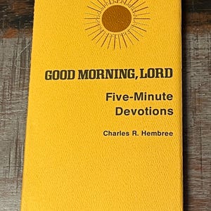 May include: A yellow book titled Good Morning, Lord Five-Minute Devotions by Charles R. Hembree. The cover features a brown sun graphic with radiating lines centered at the top. The text is printed in a bold, dark brown sans-serif font.