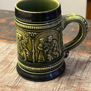 May include: A dark green ceramic beer stein featuring a relief design of figures in traditional clothing. The mug has a cylindrical body with a thick handle, a dark rim, and a dark base, showcasing detailed moulded scenes of people in motion.