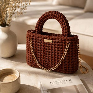 May include: A dark brown crochet handbag featuring a thick, rounded top handle and a gold-toned metal chain shoulder strap. The front of the bag is adorned with a small, rectangular gold-toned metal plaque centered near the top edge.