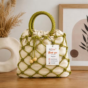 Può includere: Una borsa in tessuto color crema con un rivestimento a rete in macramè verde oliva, due manici rotondi verde oliva e due perline di legno su una coulisse. Un'etichetta rettangolare bianca attaccata alla borsa riporta il testo Timaya Thank you.
