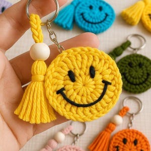 Puede incluir: Un llavero de cara sonriente de ganchillo amarillo con un cordón trenzado, una cuenta de madera natural y una borla amarilla a juego unida a una anilla de metal plateado. El diseño incluye una cara redonda de hilo amarillo con ojos y boca negros.