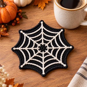 May include: A black and white crocheted coaster featuring a spider web pattern. The coaster is hexagonal in shape with a central circular point from which white web lines radiate outward to the edges, creating a geometric web design.