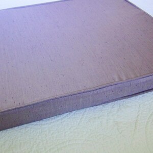 May include: A rectangular cushion with a light purple fabric cover. The cushion has a stitched edge and a subtle textured pattern.