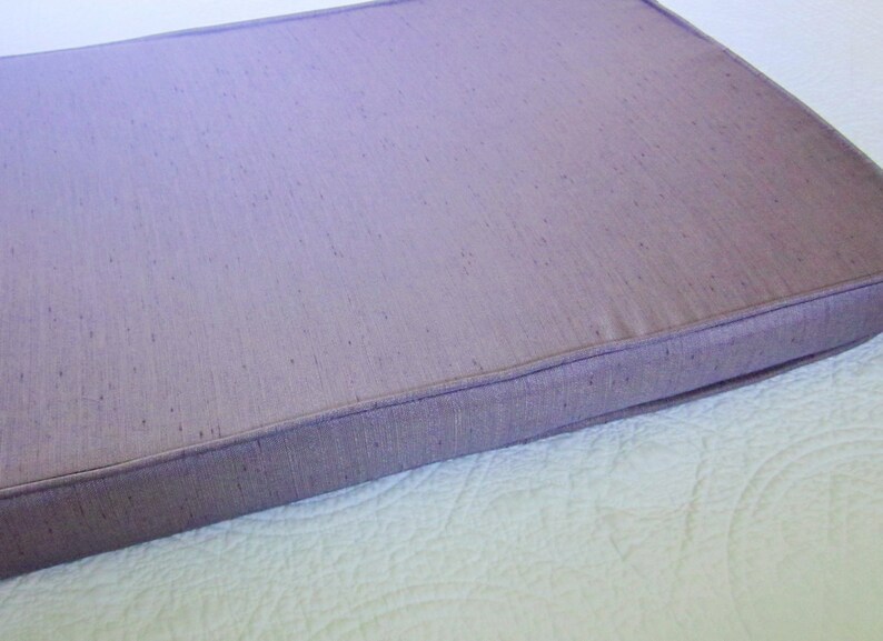 May include: A rectangular cushion with a light purple fabric cover. The cushion has a stitched edge and a subtle textured pattern.