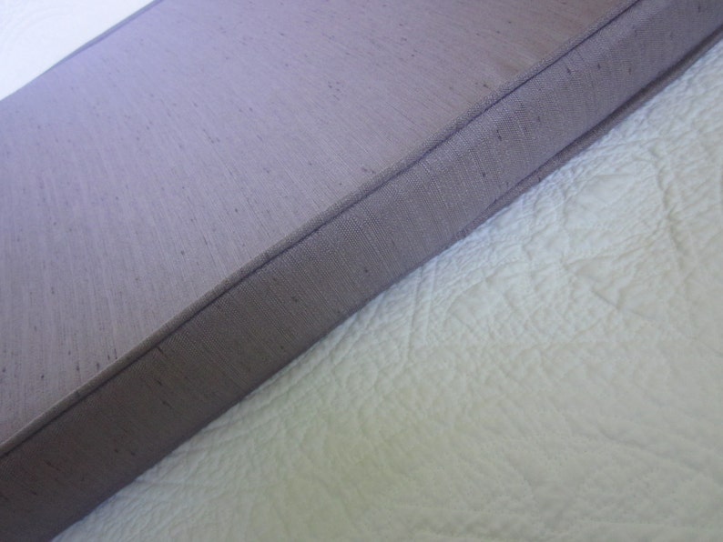 May include: A close-up of a light purple fabric with a subtle texture. The fabric has a stitched edge and is folded over a white surface.
