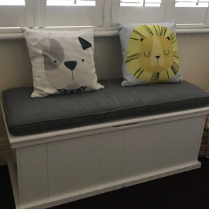 May include: White storage bench with a gray cushion and two decorative pillows. One pillow features a cartoon dog and the other a cartoon lion.