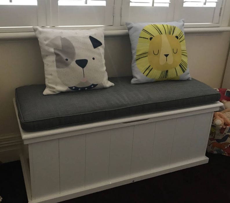 May include: White storage bench with a gray cushion and two decorative pillows. One pillow features a cartoon dog and the other a cartoon lion.
