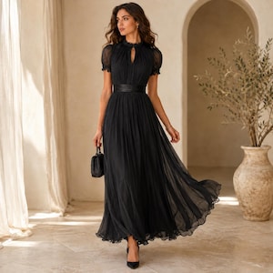 May include: A floor-length black dress featuring short puff sleeves, a high neckline with a keyhole cutout, and a fitted waist with a wide band. The skirt is pleated and flows to the floor, made of a sheer, layered fabric. A small black handbag is held.