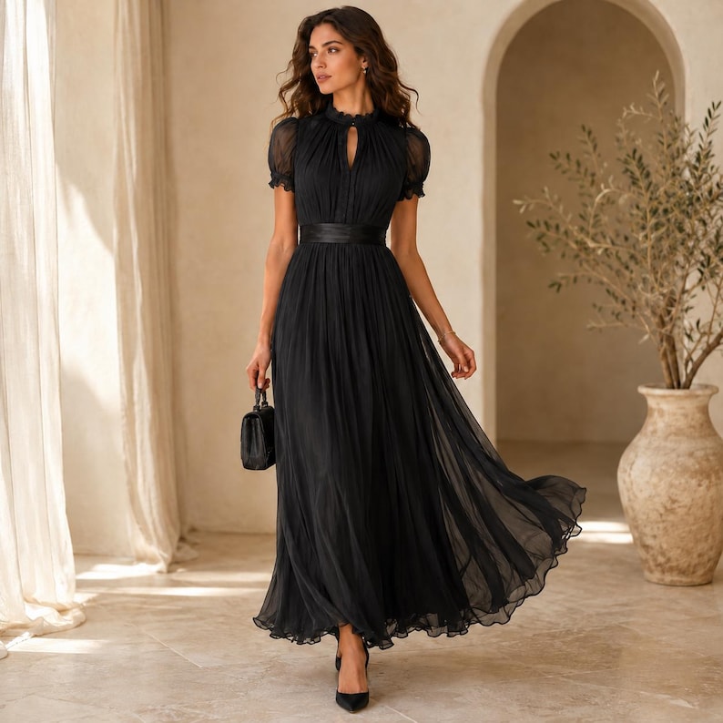 May include: A floor-length black dress featuring short puff sleeves, a high neckline with a keyhole cutout, and a fitted waist with a wide band. The skirt is pleated and flows to the floor, made of a sheer, layered fabric. A small black handbag is held.