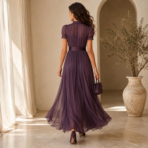 May include: A floor-length deep purple dress featuring a high neckline, short puffed sleeves, and a fitted waist with a wide band. The skirt is made of pleated, sheer fabric that flows to the floor, and the garment is paired with a matching handbag.