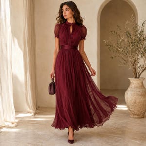 May include: A floor-length burgundy dress featuring a high neckline, short puff sleeves, and a fitted waist with a wide band. The skirt is made of a sheer, pleated fabric that flows to the floor. A small, matching burgundy handbag is held in one hand.
