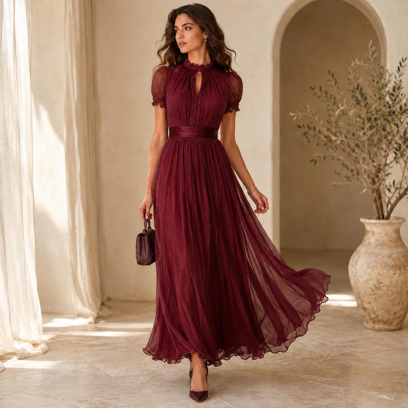 May include: A floor-length burgundy dress featuring a high neckline, short puff sleeves, and a fitted waist with a wide band. The skirt is made of a sheer, pleated fabric that flows to the floor. A small, matching burgundy handbag is held in one hand.