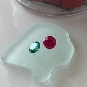 May include: A faceted oval-shaped green gemstone and a faceted round-shaped red gemstone resting on a smooth, translucent, pale seafoam green piece of sea glass. The gemstones exhibit clear, reflective surfaces with multiple cut facets.