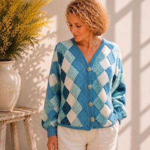 Könnte beinhalten: Blau, weiß und hellblau gemusterte Strickjacke mit V-Ausschnitt und Knopfverschluss. Die Strickjacke hat lange Ärmel und wird mit einer weißen Hose getragen. Eine Vase mit gelben Blumen steht auf einem Holzschemel.