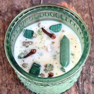 May include: A white wax candle in a textured green glass jar, topped with green crystals, a double-terminated green stone point, and dried brown cloves. The candle surface is sprinkled with small green and metallic gold-colored mineral fragments.