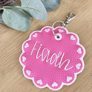 May include: A circular pink keyring with a scalloped edge featuring embroidered light pink hearts along the border. The centre displays the name Fiaah in white cursive embroidery. A silver metal split ring and lobster clasp are attached.