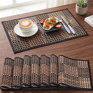 May include: A set of rectangular placemats featuring a woven bamboo pattern in alternating light tan and black squares with a solid black fabric border. The placemats are shown both laid flat with a coffee cup and plate and stacked in a folded row.