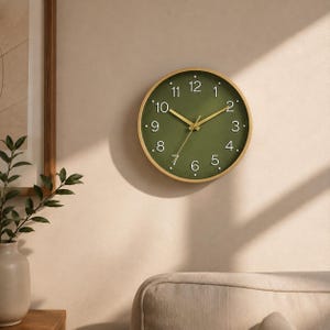 Puede incluir: Un reloj de pared redondo con marco de madera clara y esfera de color verde oliva mate. El reloj presenta números arábigos blancos del 1 al 12, manecillas de hora y minuto doradas finas y un segundero dorado fino en el centro.