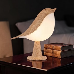 May include: A bird-shaped table lamp featuring a light wood grain finish on the head, tail, and base, with a smooth, glowing white translucent body that emits a soft light. The lamp stands on a circular base next to two stacked antique books.