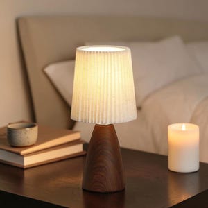 May include: A table lamp featuring a conical dark brown wooden base and a white pleated fabric lampshade. The lamp emits a warm glow, illuminating the surrounding area with a soft light, creating a focused and clear visual presence on the surface.
