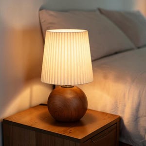 May include: A table lamp featuring a spherical wooden base with a natural grain finish and a white, vertically pleated, tapered fabric lampshade. The lamp emits a warm, soft glow, illuminating the surrounding area with a gentle light.