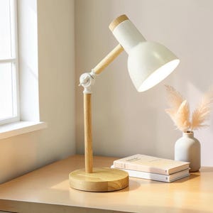May include: A desk lamp featuring a light-coloured wooden base, a vertical wooden stem, and a white metal shade. The lamp has an adjustable arm with a white plastic hinge and a wooden accent piece connecting the shade to the main structure.