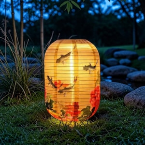 May include: A cylindrical paper lantern with a warm internal glow, featuring a printed design of dark koi fish swimming among vibrant red and green floral motifs. The lantern has a ribbed structure and sits on a patch of green grass.